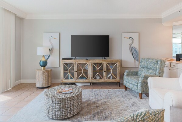 Una sala de estar con televisor, un aparador con puertas de cristal, un sillón estampado, un reposapiés redondo y una lámpara.