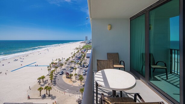 Un balcon avec une table et des chaises, donnant sur une plage bordée de palmiers et une route.