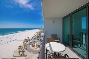 Un balcón con mesa y sillas, con vista a una playa con palmeras y una carretera.