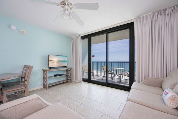 A room with a TV, a dining table with chairs, a sofa, and a balcony with a view of the sea.