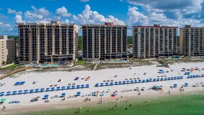 Un hôtel en bord de mer composé de plusieurs immeubles de grande hauteur, une plage de sable fin avec des parasols et des chaises longues, et un ciel bleu dégagé.