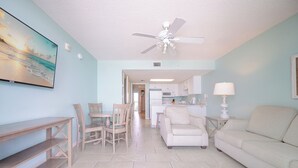 A living room with a ceiling fan, a dining area with chairs, a sofa, a TV, and a kitchen in the background.