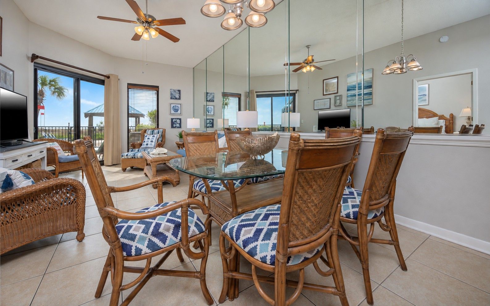A spacious living area with a glass-top dining table surrounded by wicker chairs with patterned cushions, a ceiling fan, and a large mirror reflecting the room.