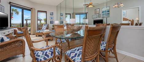 A spacious living area with a glass-top dining table surrounded by wicker chairs with patterned cushions, a ceiling fan, and a large mirror reflecting the room.