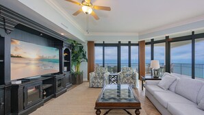Ein Wohnzimmer mit großem Flachbildfernseher, Deckenventilator, Sofa, Couchtisch und Meerblick durch große Fenster.
