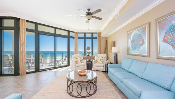 A spacious living room with a large window overlooking the beach, a ceiling fan, and a blue sofa. - The Oasis at Orange Beach, formerly known as Phoenix West II, Unit 303 (Orange Beach)
