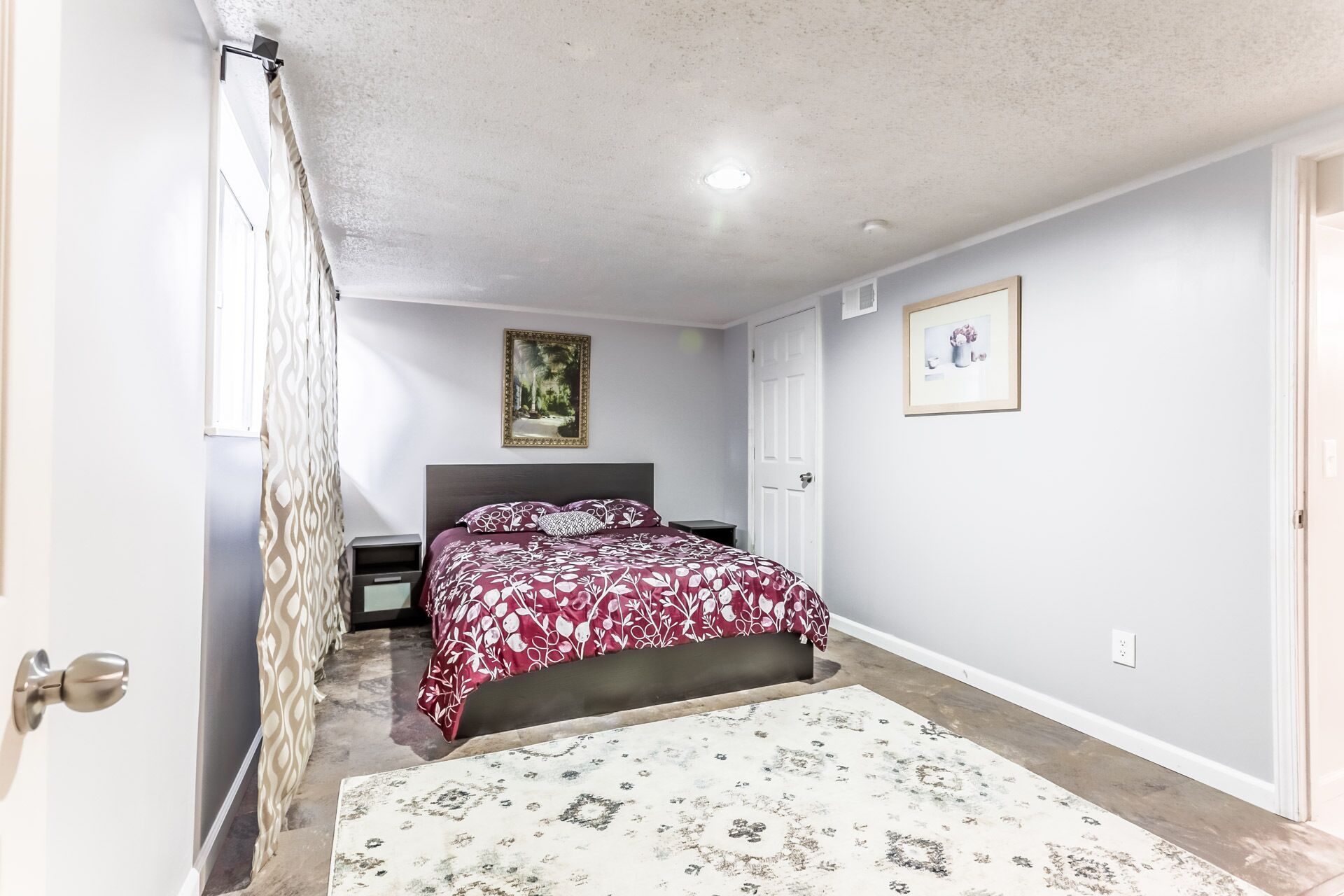 A bedroom with a bed, a nightstand, a door, a framed picture, and a patterned rug.