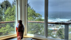 Una ventana con vista al océano y árboles, un florero y un libro sobre una mesa.