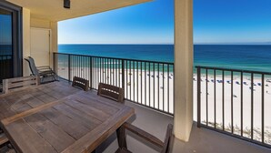 Un balcon avec une table et des chaises en bois, donnant sur une plage parsemée de parasols et fréquentée par des gens.