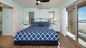 A bedroom with a bed, a ceiling fan, two bedside tables with lamps, a door, and a large window with a view of the beach.