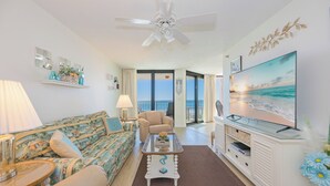 Un salon comprenant un canapé, une table basse et un téléviseur. On y trouve également un ventilateur de plafond, un balcon avec vue et un élément décoratif mural.