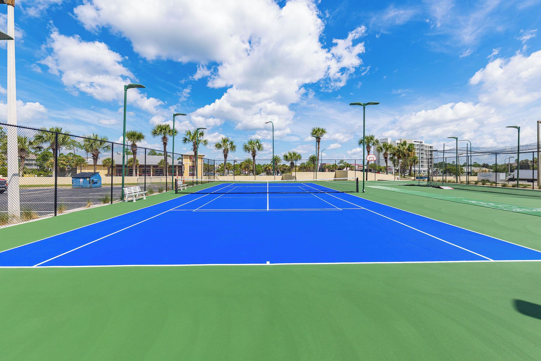 Un court de tennis avec un filet, entouré d’une clôture, de palmiers et de bâtiments en arrière-plan.