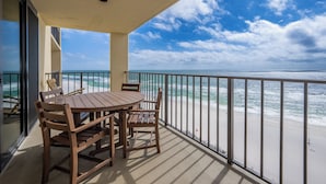 Uma varanda com mesa e cadeiras de madeira, com vista para uma praia com ondas e céu claro.