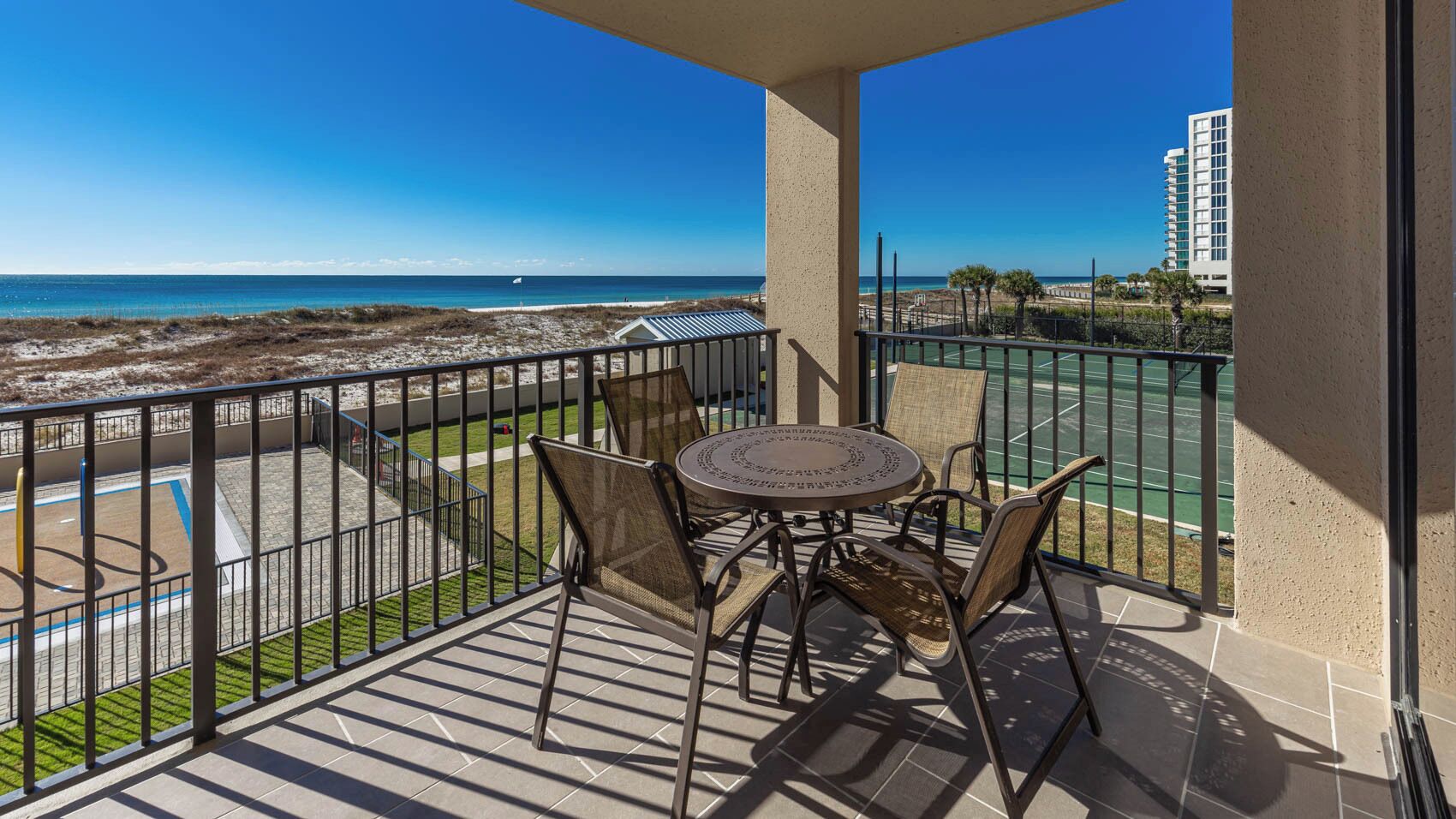 A balcony with a view of the beach, a tennis court, and a building in the background.