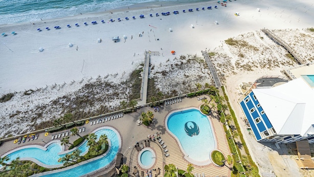 Vista aérea de un complejo turístico con zona de piscina, sombrillas de playa y un paseo marítimo que conduce a la playa.