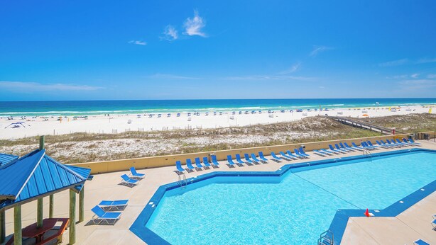 A pool with lounge chairs, a beach view, and a clear sky.