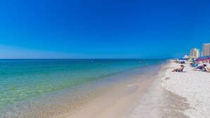A beach with clear turquoise water, white sand, and a few people sunbathing. There are umbrellas and beach chairs along the shore. - SUNSET VIEW ~ Balcony on the BEACH! ~ Phoenix Hotels (Gulf Shores)