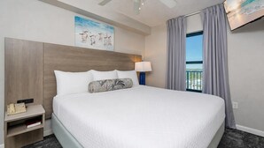 A hotel room with a large bed, a ceiling fan, a painting on the wall, a TV, and a window with curtains. - SUNSET VIEW ~ Balcony on the BEACH! ~ Phoenix Hotels (Gulf Shores)
