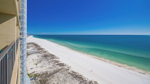 Une vue sur la plage depuis un balcon, avec un ciel bleu dégagé, une plage de sable et l’océan.