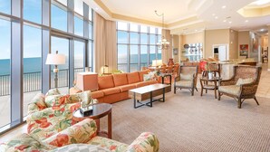 A spacious living area with a large window overlooking the ocean, a cozy seating arrangement, and a kitchen in the background. - The Oasis at Orange Beach, formerly known as Phoenix West II, Unit 201 (Orange Beach)