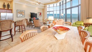 A spacious living room with a large dining table, wicker chairs, and a view of the ocean through large windows. - The Oasis at Orange Beach, formerly known as Phoenix West II, Unit 201 (Orange Beach)