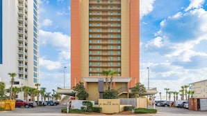 A multi-story hotel with a central entrance, balconies, and a parking area with cars and palm trees.