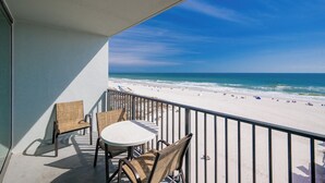 A balcony with wicker chairs and a table, overlooking a beach with people and the ocean.