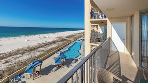 A beach view from a balcony with a swimming pool and lounge chairs.