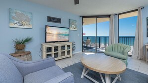 A living room with a TV, a sofa, a round table, and a balcony with a view of the sea.