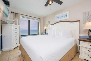 A large bed with a white headboard, a nightstand with a lamp, a dresser, a ceiling fan, and a window with a view of the ocean.