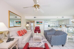 A living room with a sofa, armchair, and a coffee table. There is a kitchen in the background with a microwave and a refrigerator.