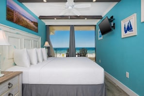 A bedroom with a large bed, a nightstand, a lamp, a TV, and a view of the beach.