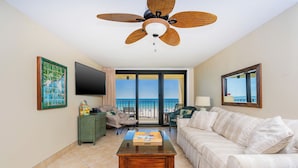 A living room with a sofa, coffee table, and a ceiling fan. There is a TV on the wall and a window with a view of the beach. - **3rd Floor ~ 2 Bedroom at Phoenix II** (Orange Beach)