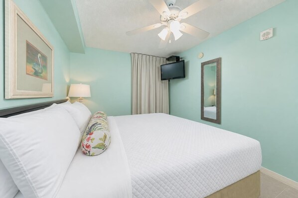 Habitación de hotel con una cama, ventilador de techo, televisor, espejo y un cuadro en la pared.