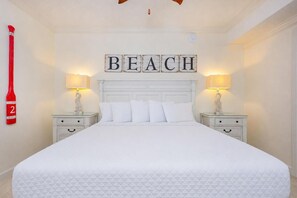 A hotel room with a large bed, two bedside tables with lamps, a wall-mounted sign reading 'BEACH', and a red object on the wall.