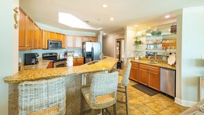 A kitchen with wooden cabinets, a granite countertop, a built-in microwave, a refrigerator, and a glass-fronted display shelf. - Phoenix West 305 Your Coastal Getaway! (Orange Beach)