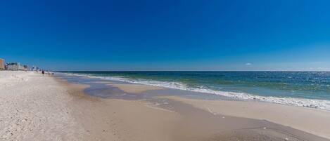 Une vaste plage de sable fin, avec des vagues douces, un ciel bleu clair et des bâtiments en arrière-plan.