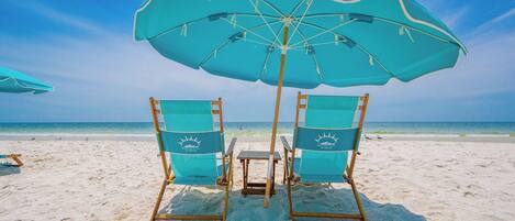 Two beach chairs with a table under a large umbrella on a sandy beach.
