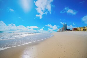 Ein Strand mit blauem Himmel, Meereswellen und Gebäuden im Hintergrund.