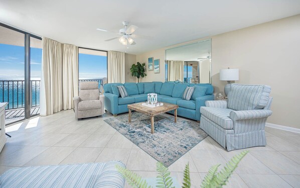 A spacious living room with a large blue sofa, a beige armchair, and a striped armchair. A wooden coffee table is in the center, and a large window offers a view of the ocean.