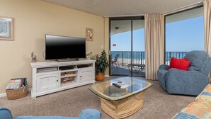 Ein Wohnzimmer mit Flachbildfernseher auf einer weißen Unterhaltungseinheit, einem Glastisch, einem blauen Sofa mit rotem Kissen und Blick auf den Strand durch eine Glastür.