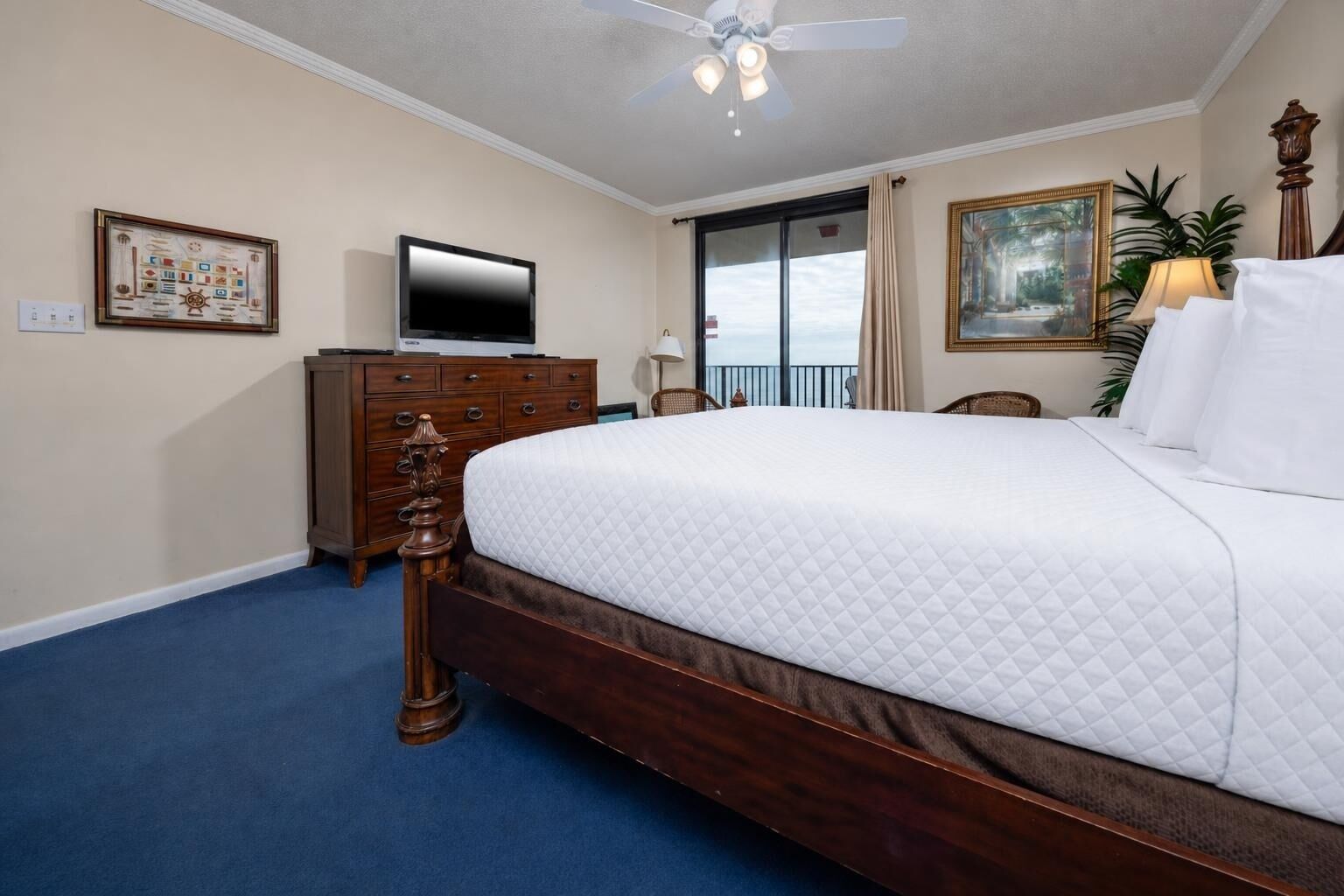 A hotel room with a large bed, a dresser, a television, a ceiling fan, and a painting on the wall.