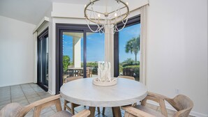 A dining area with a round table, wooden chairs, a chandelier, and large sliding glass doors leading to an outdoor patio with palm trees. - GROUND FLOOR! 1 BR Condo in High Demand! (Orange Beach)