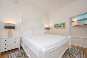 Un lit à baldaquin blanc avec un matelas matelassé, une table de chevet avec une lampe, un tableau représentant des chaises de plage et un téléviseur fixé au mur.