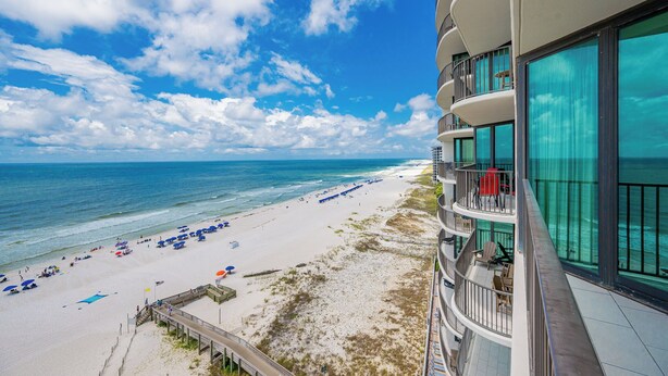 A beachfront hotel with a balcony overlooking the ocean, a sandy beach with umbrellas, and a clear sky. - ~Beach Front~ 2 Bedroom @ Phoenix IX (Orange Beach)