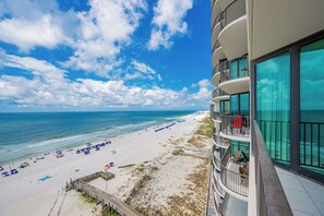 A beachfront hotel with a balcony overlooking the ocean, a sandy beach with umbrellas, and a clear sky. - ~Beach Front~ 2 Bedroom @ Phoenix IX (Orange Beach)