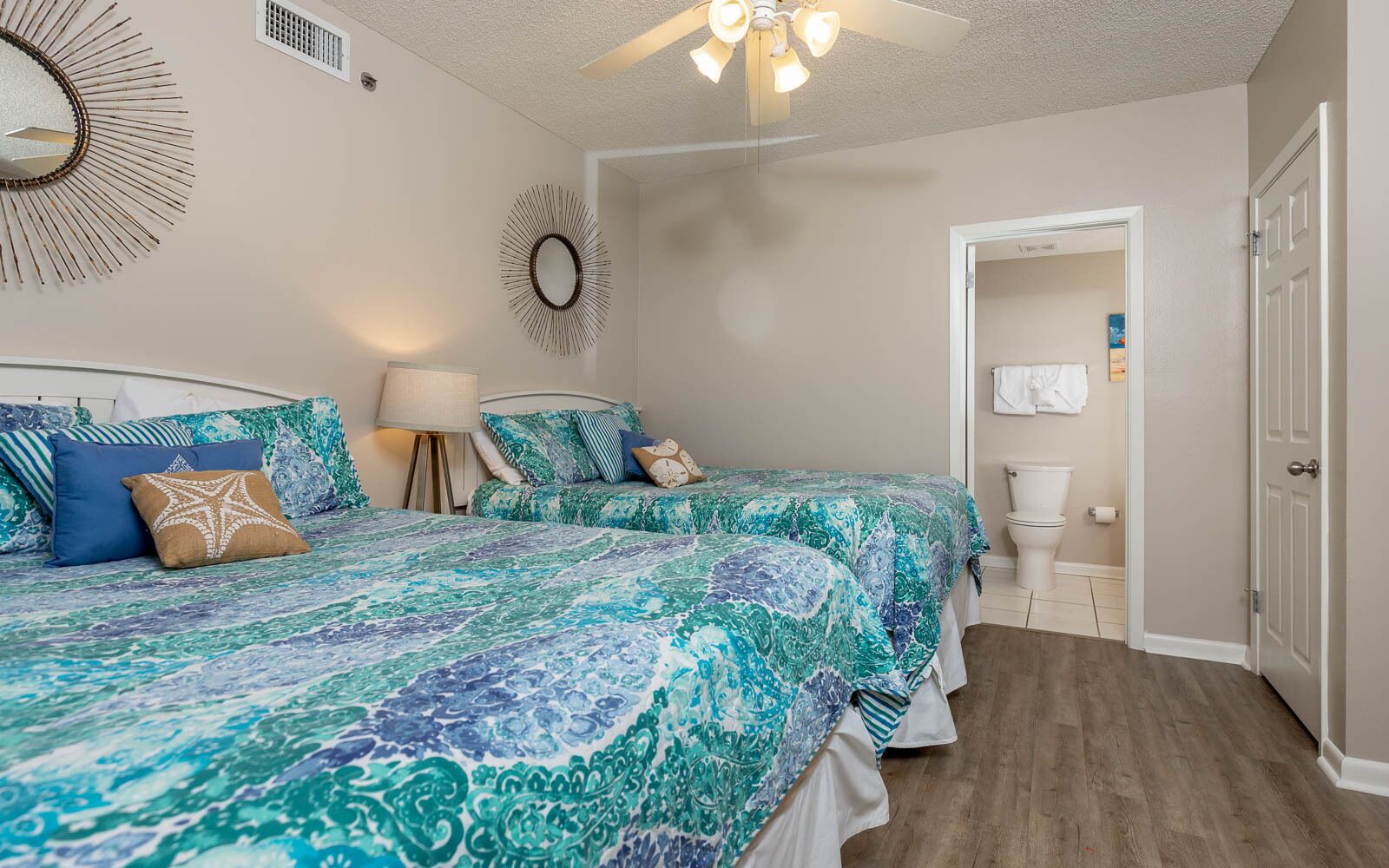 A bedroom with two beds, a ceiling fan, a wall mirror, and a bathroom visible through an open door.