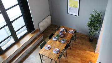 Uma área de jantar com uma mesa de madeira posta para quatro, cadeiras, um radiador, um quadro na parede e um vaso com planta.