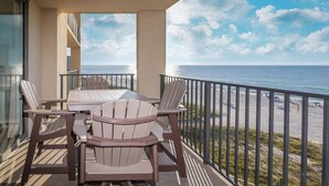 A balcony with Adirondack chairs and a table, overlooking a beach with a clear sky.