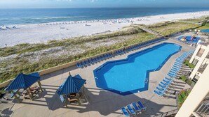 A swimming pool with blue chairs and umbrellas, a beach with people, and a view of the ocean.